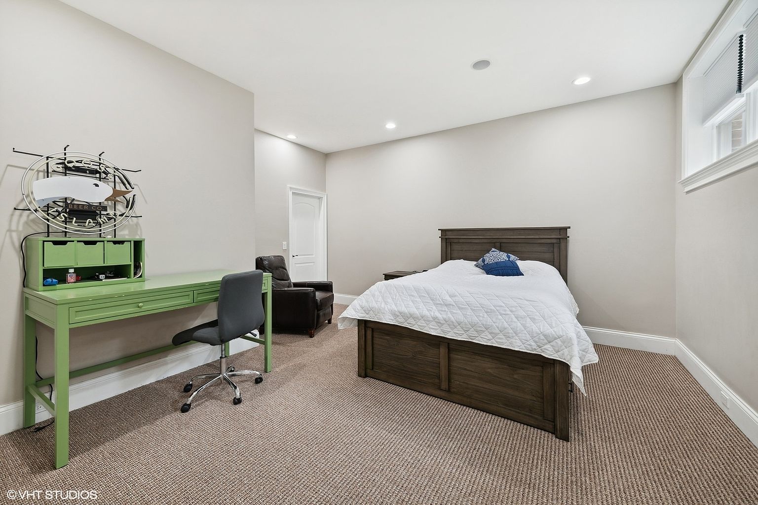 This is a well-lit guest bedroom featuring a wooden bed with a white comforter and a blue decorative pillow. The room includes a green desk with a gray chair, and a leather armchair, suggesting a combined sleeping and study area. The carpeted floor and neutral wall colors create a cozy and inviting atmosphere.