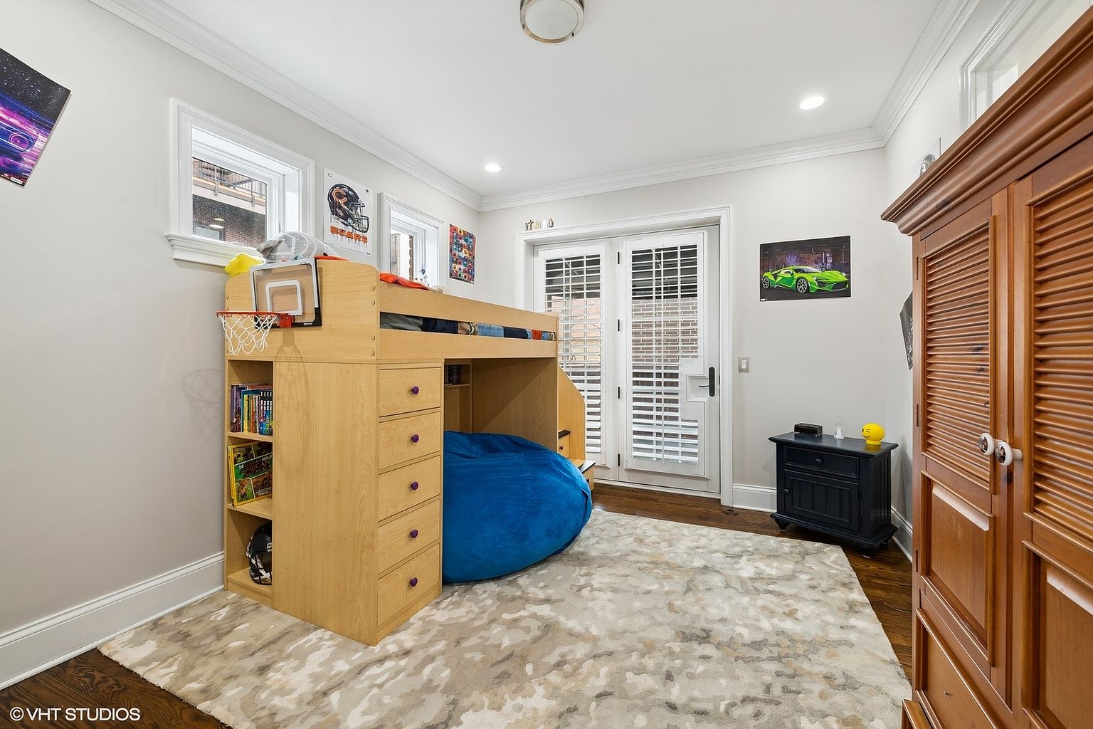 This is a well-lit bedroom featuring a unique loft bed with built-in storage and a basketball hoop. A large blue beanbag chair sits beneath the loft, adding a playful touch. The room is decorated with sports-themed artwork and has a neutral-toned rug, creating a comfortable and inviting space.