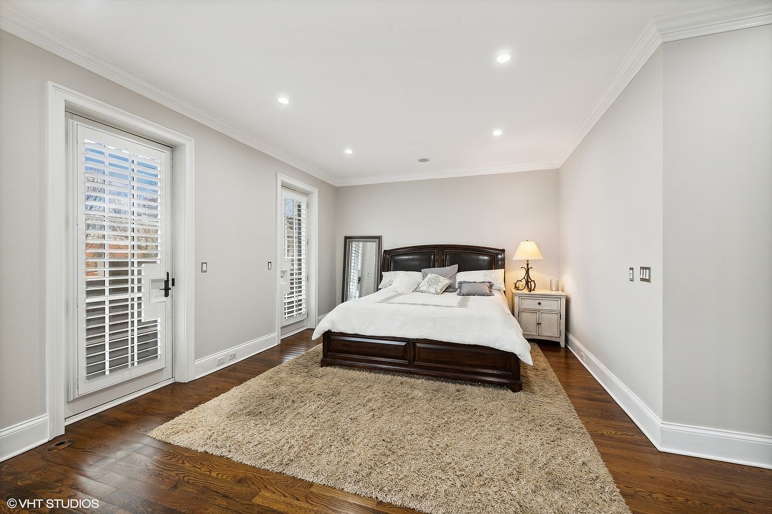 This is a well-lit primary bedroom featuring a dark wood bed frame with white linens and decorative pillows, set upon a large, neutral-toned shag rug. The room has hardwood floors, light gray walls, white trim, and recessed lighting. A full-length mirror and a bedside table with a lamp add to the room's functionality and aesthetic appeal.