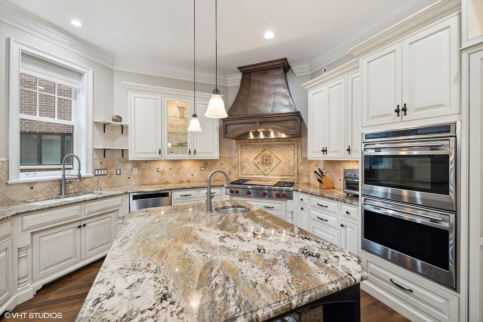 This is a well-lit kitchen featuring white cabinetry, stainless steel appliances, and a large island with a granite countertop. The kitchen also has a decorative backsplash and a custom range hood. The perspective is from the center of the kitchen, showcasing the island and the surrounding cabinetry.