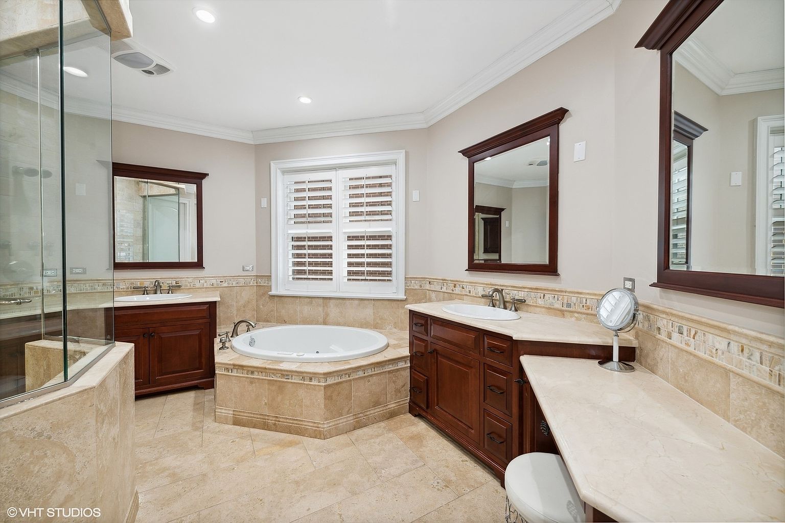 This is a primary bathroom featuring a large soaking tub surrounded by beige tile, a separate glass-enclosed shower, and dual vanities with dark wood cabinets and light countertops. The room is well-lit with recessed lighting and natural light from a window with shutters. The overall style is luxurious and traditional.