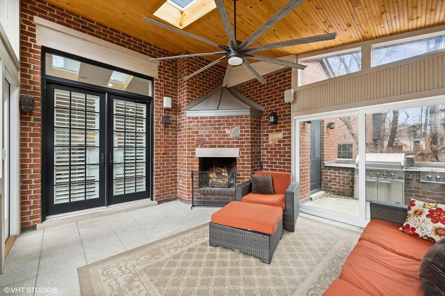 This is an inviting sunroom featuring a brick fireplace, a ceiling fan, and comfortable seating. The room is well-lit with natural light from the windows and skylight. The space is designed for relaxation and enjoyment of the outdoors, with a seamless transition to an outdoor patio area.