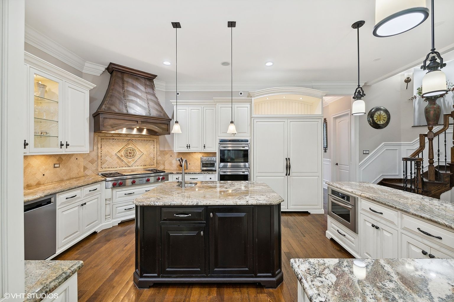 This is a well-appointed kitchen featuring custom cabinetry, granite countertops, and stainless steel appliances. A large island with a dark base provides ample workspace and seating. The kitchen also includes a decorative range hood and a staircase leading to the upper level, creating a luxurious and inviting space.