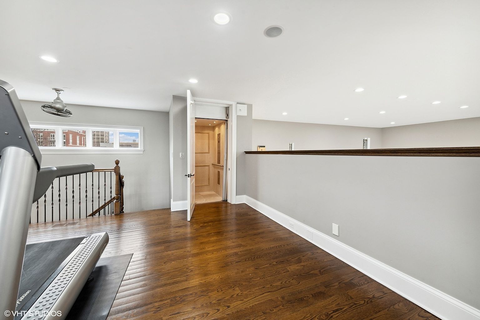 This interior shot showcases a home gym area with hardwood floors and light gray walls. An open elevator is visible, suggesting accessibility, while a treadmill is positioned in the foreground. The space is well-lit with recessed lighting, creating a clean and inviting atmosphere.
