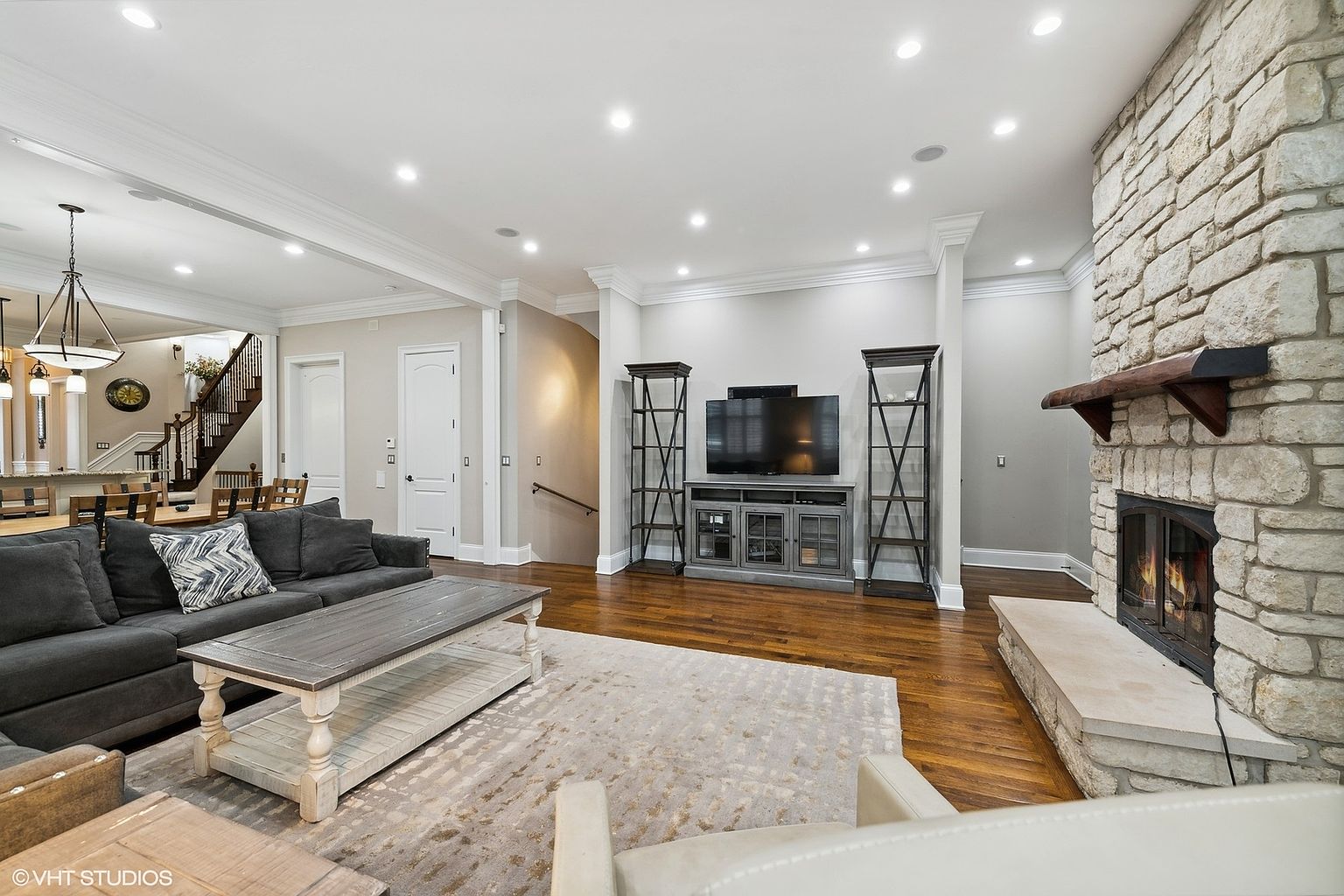 This is an interior shot of a living room featuring a stone fireplace, a dark gray sectional sofa, and a light-colored area rug. The room has hardwood floors and is well-lit with recessed lighting. A television is mounted above a console table, flanked by decorative shelving units.