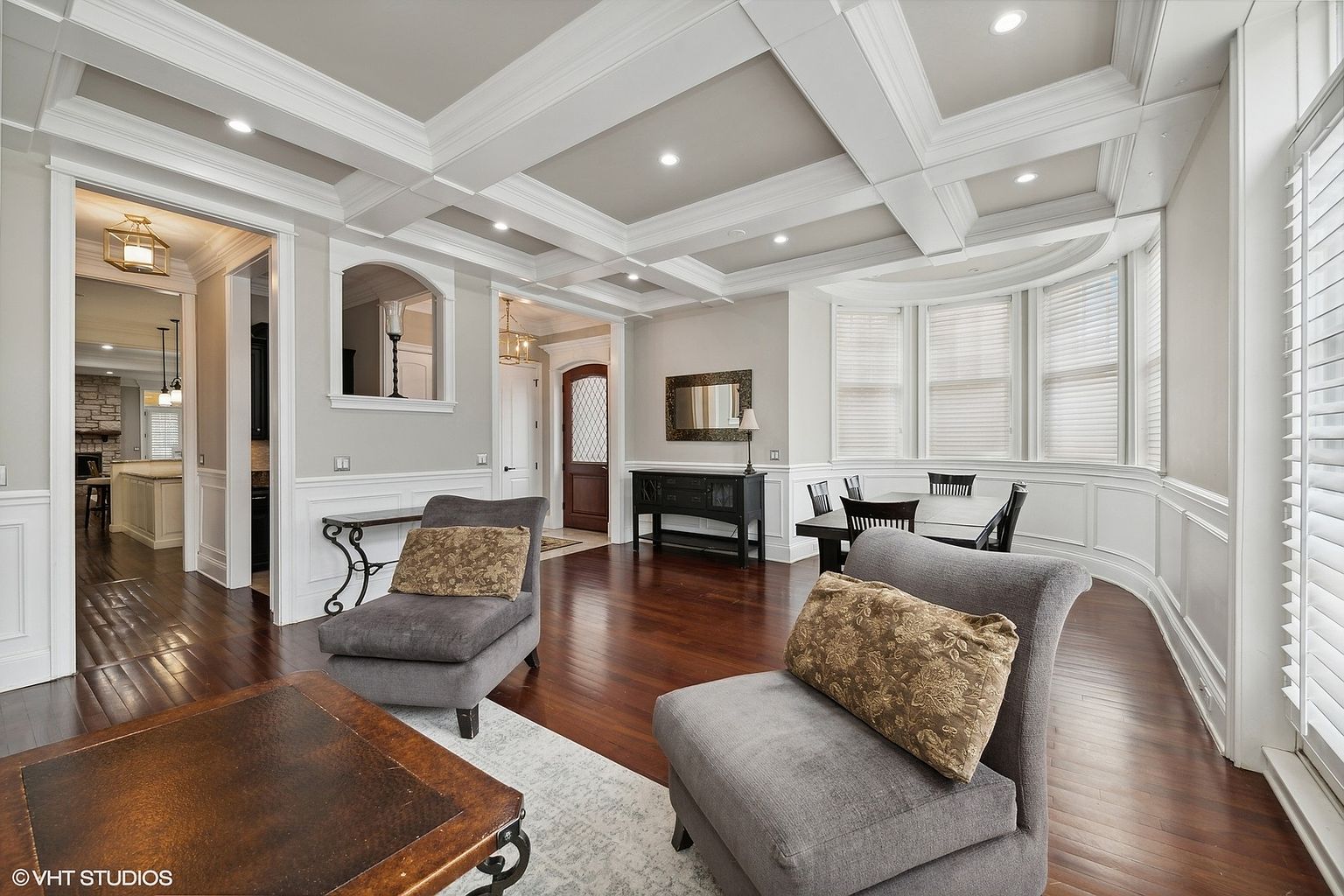 This is an interior shot of a living room featuring hardwood floors, coffered ceilings, and a bay window. Two gray armchairs with decorative pillows are positioned in the foreground, and a dark wood console table with a mirror sits against the wall. The room exudes a sense of classic elegance and comfort.