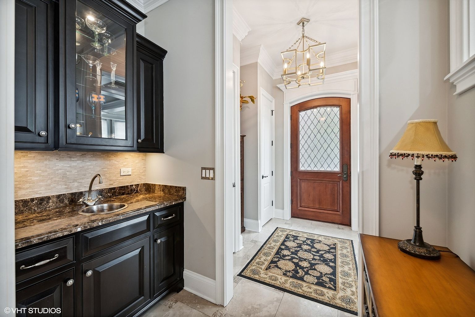 This interior shot showcases a hallway featuring a dark wood door with a diamond-patterned glass insert, illuminated by a modern chandelier. To the left, a built-in bar area with dark cabinetry and a marble countertop adds a touch of luxury. The hallway is decorated with a patterned rug, and a lamp sits on a wooden console table to the right, creating a warm and inviting atmosphere.