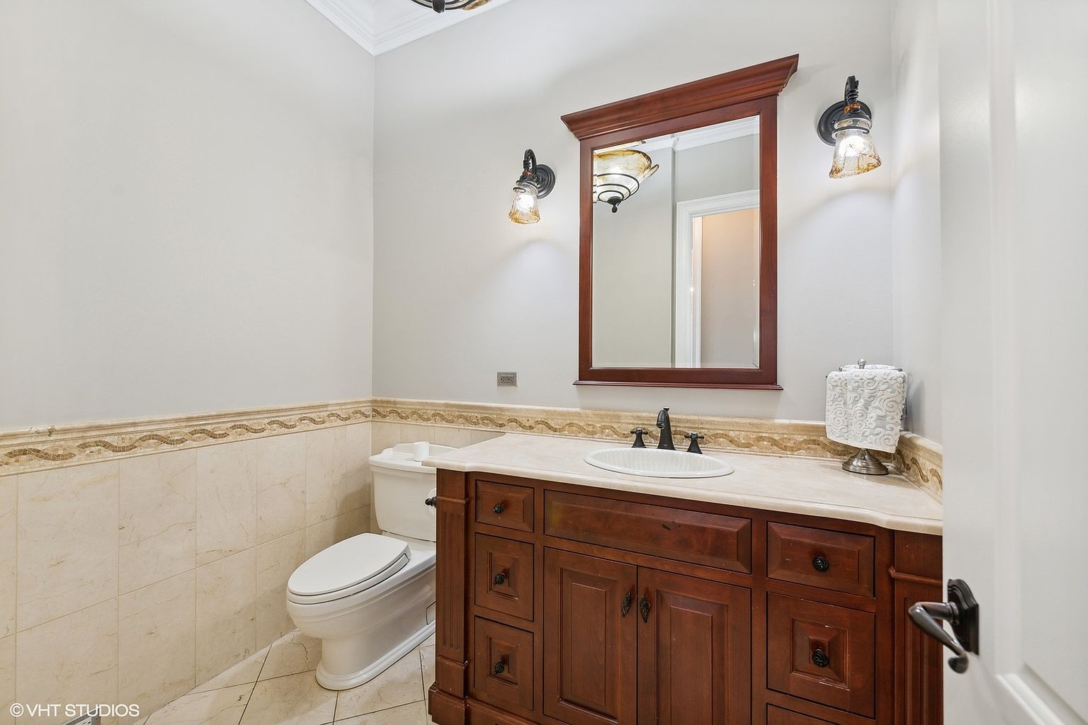 This is a well-maintained guest bathroom featuring a dark wood vanity with a light-colored countertop and an oval sink. A framed mirror hangs above the vanity, flanked by decorative sconces. The toilet is positioned to the left, and the walls are adorned with tile and a decorative border, creating a classic and clean aesthetic.