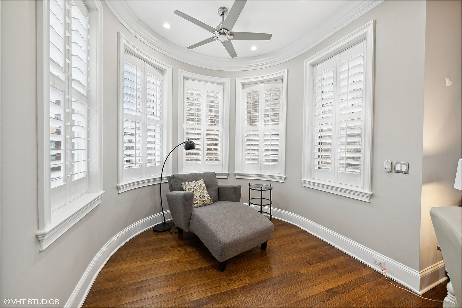 This is a bright and inviting sunroom featuring a curved wall with large windows fitted with white shutters, allowing ample natural light. The room is furnished with a comfortable gray chaise lounge and a small side table, set against a backdrop of rich hardwood flooring. A ceiling fan provides air circulation, enhancing the room's appeal as a relaxing retreat.