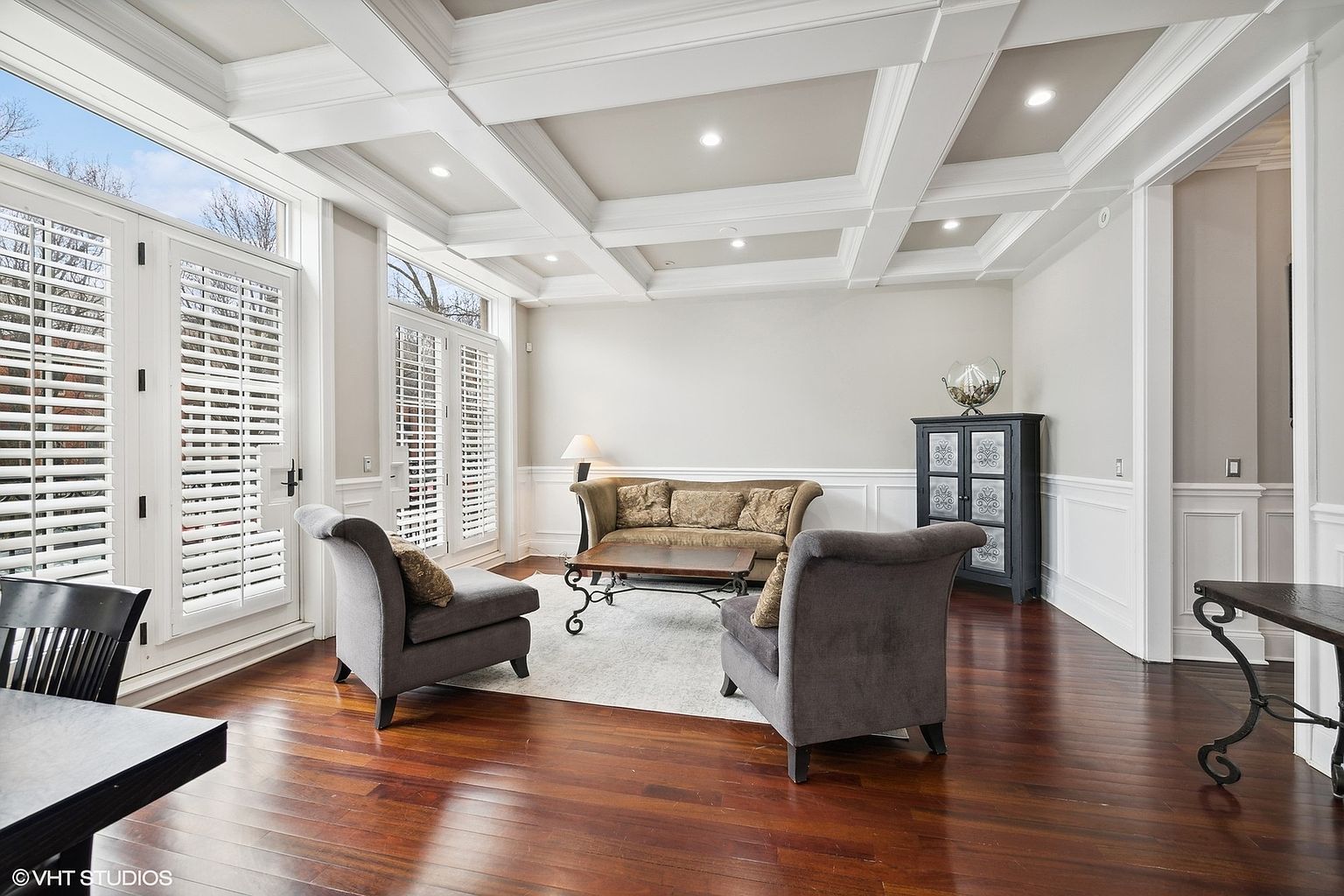 This is an interior shot of a living room featuring hardwood floors, a coffered ceiling, and white trim. The room is furnished with a sofa, two armchairs, a coffee table, and a dark cabinet. Natural light streams in through the shuttered windows, creating a bright and inviting atmosphere.