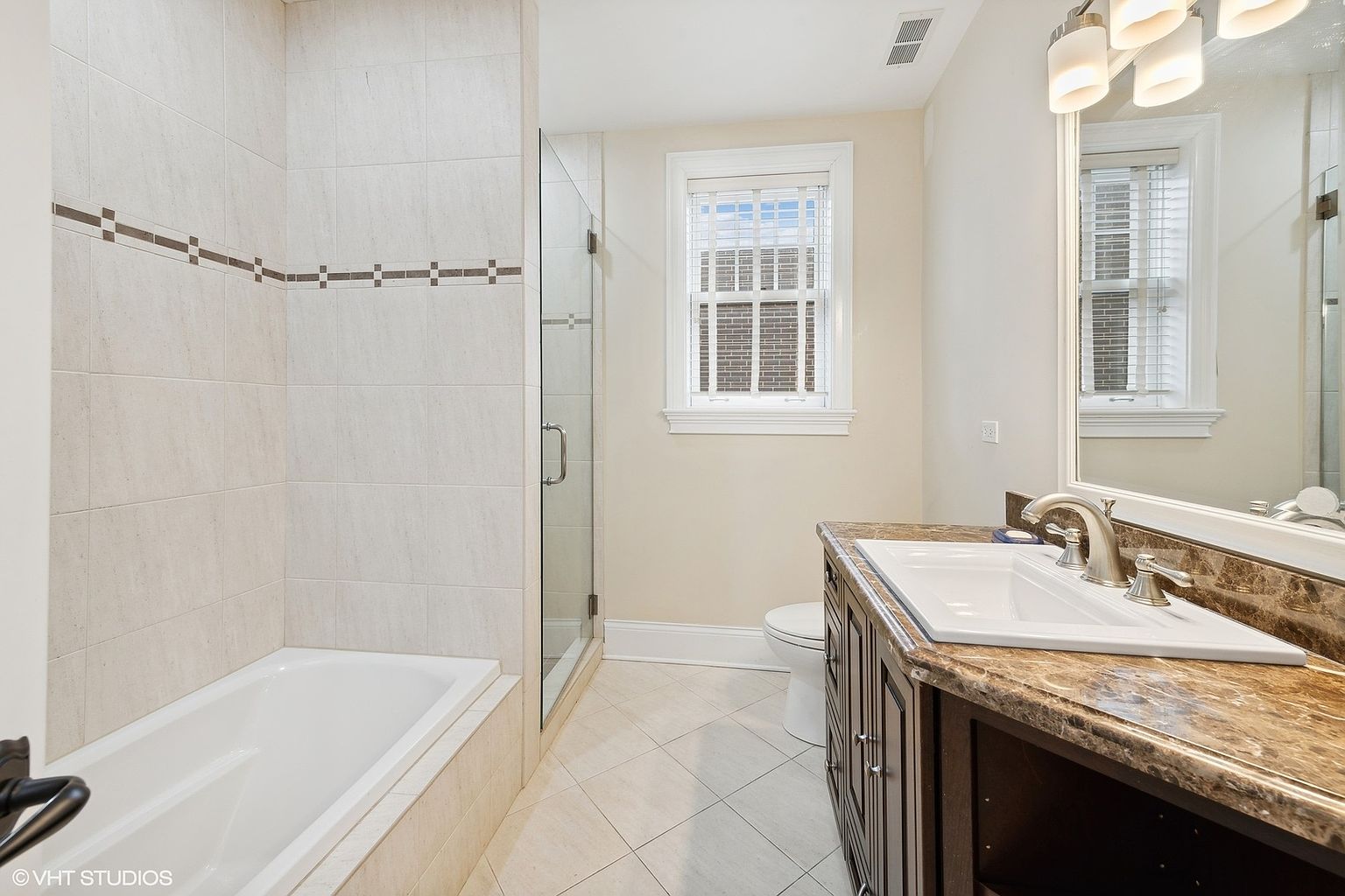 This is a well-lit bathroom featuring a bathtub with tiled walls and a decorative accent stripe, a glass-enclosed shower, and a vanity with a marble countertop. The room has a neutral color palette with beige tiles and walls, creating a clean and inviting space. A window with blinds provides natural light, and the overall impression is one of functionality and comfort.