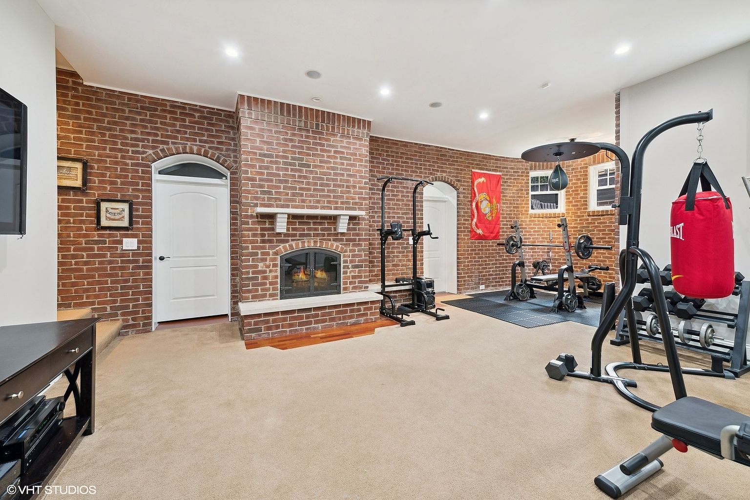 This is an interior shot of a home gym featuring exposed brick walls and a fireplace. The room is equipped with various weightlifting machines, a punching bag, and free weights. The flooring is carpeted, and the overall impression is a dedicated and well-equipped workout space.
