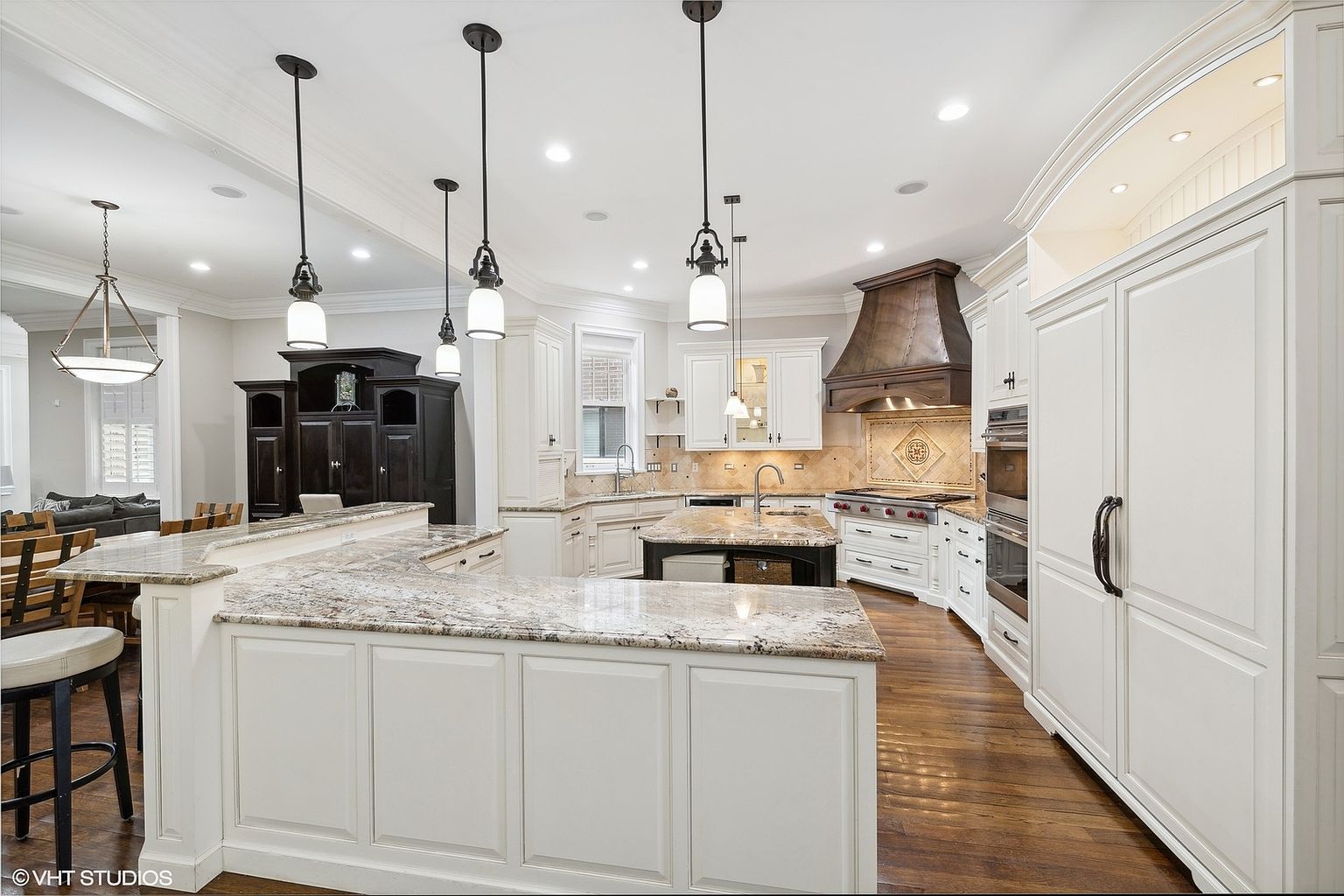 This is a bright and spacious kitchen featuring white cabinetry, granite countertops, and stainless steel appliances. A large island provides ample workspace and seating, while pendant lights add a touch of elegance. The kitchen also includes a range hood and a built-in refrigerator, creating a functional and stylish space.