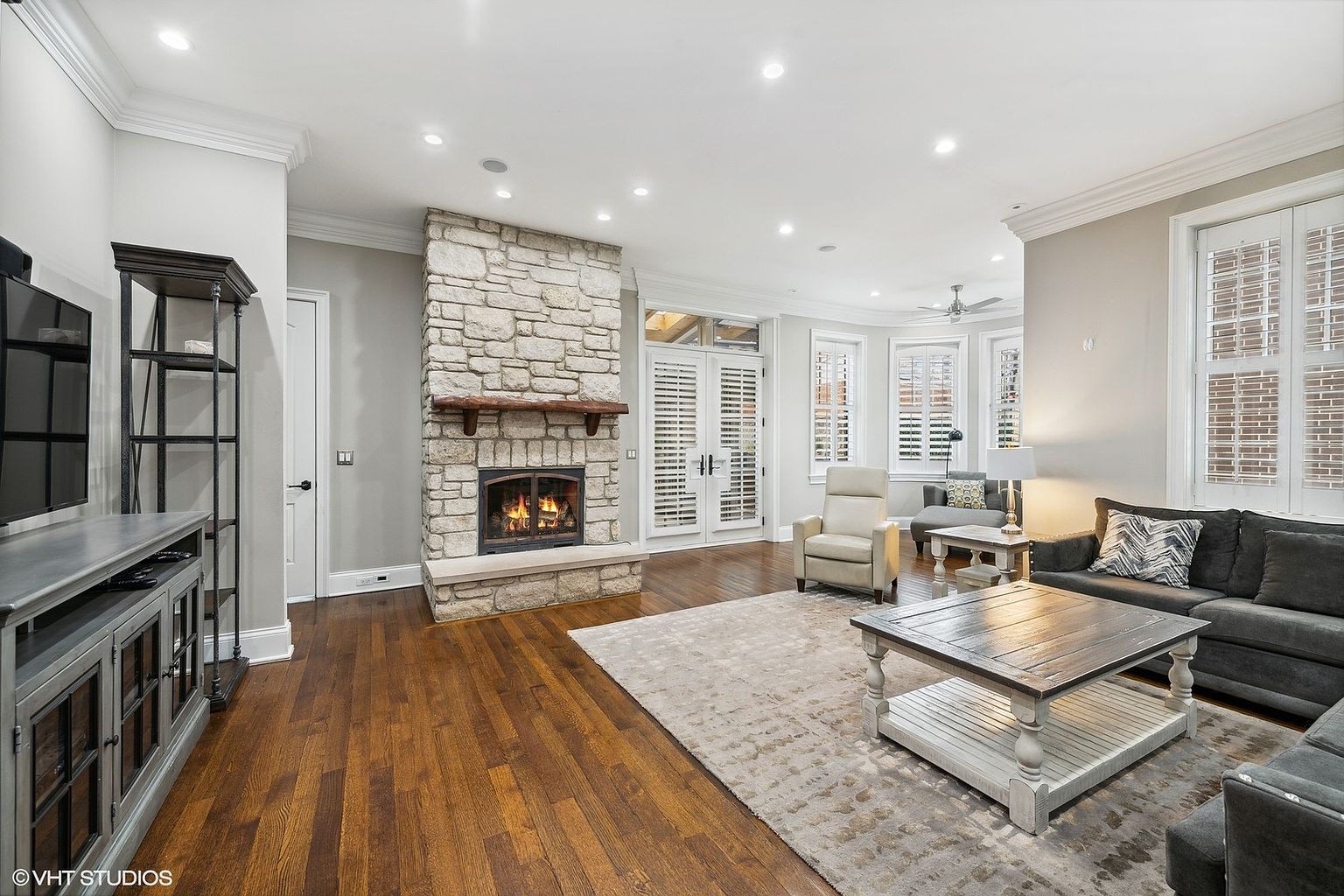 This is a well-lit living room featuring hardwood floors, a stone fireplace, and neutral-toned walls. The room is furnished with a gray sofa, a beige armchair, and a light-colored area rug, creating a comfortable and inviting atmosphere. French doors with white shutters provide access to an outdoor area, enhancing the room's appeal.