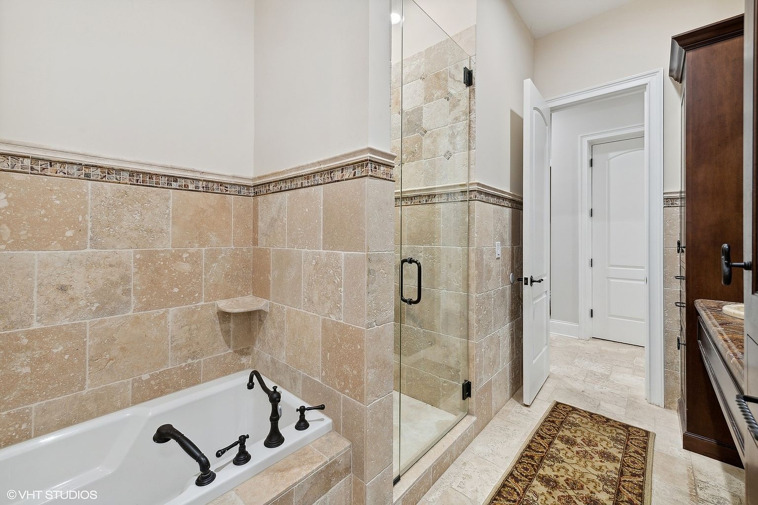This is a well-appointed primary bathroom featuring travertine tile walls and flooring, a soaking tub with black fixtures, and a glass-enclosed shower. The bathroom also includes a dark wood vanity with a marble countertop and an adjacent doorway leading to another room. The overall impression is luxurious and spa-like.