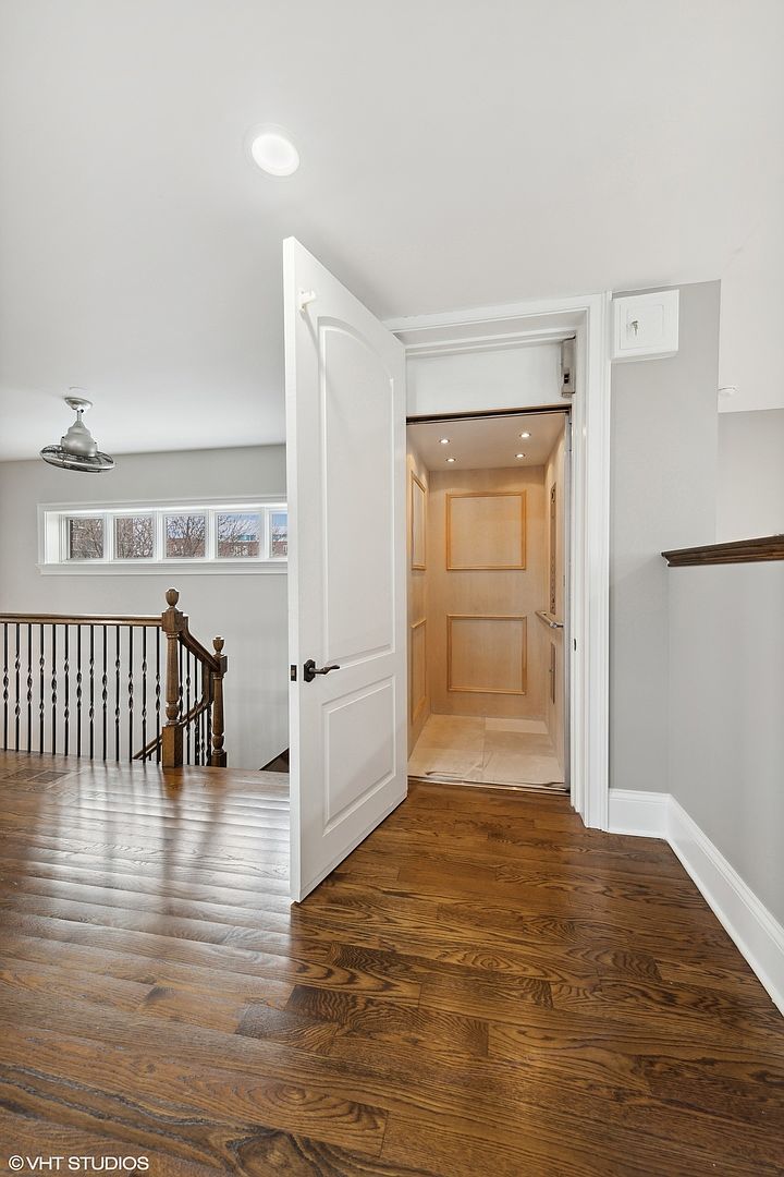This interior shot showcases a hallway with dark hardwood flooring and a staircase leading down. An open white door reveals an elevator with light wood paneling, adding a unique feature to the home. The walls are painted in a neutral gray, and natural light filters in through a window, creating a bright and inviting space.
