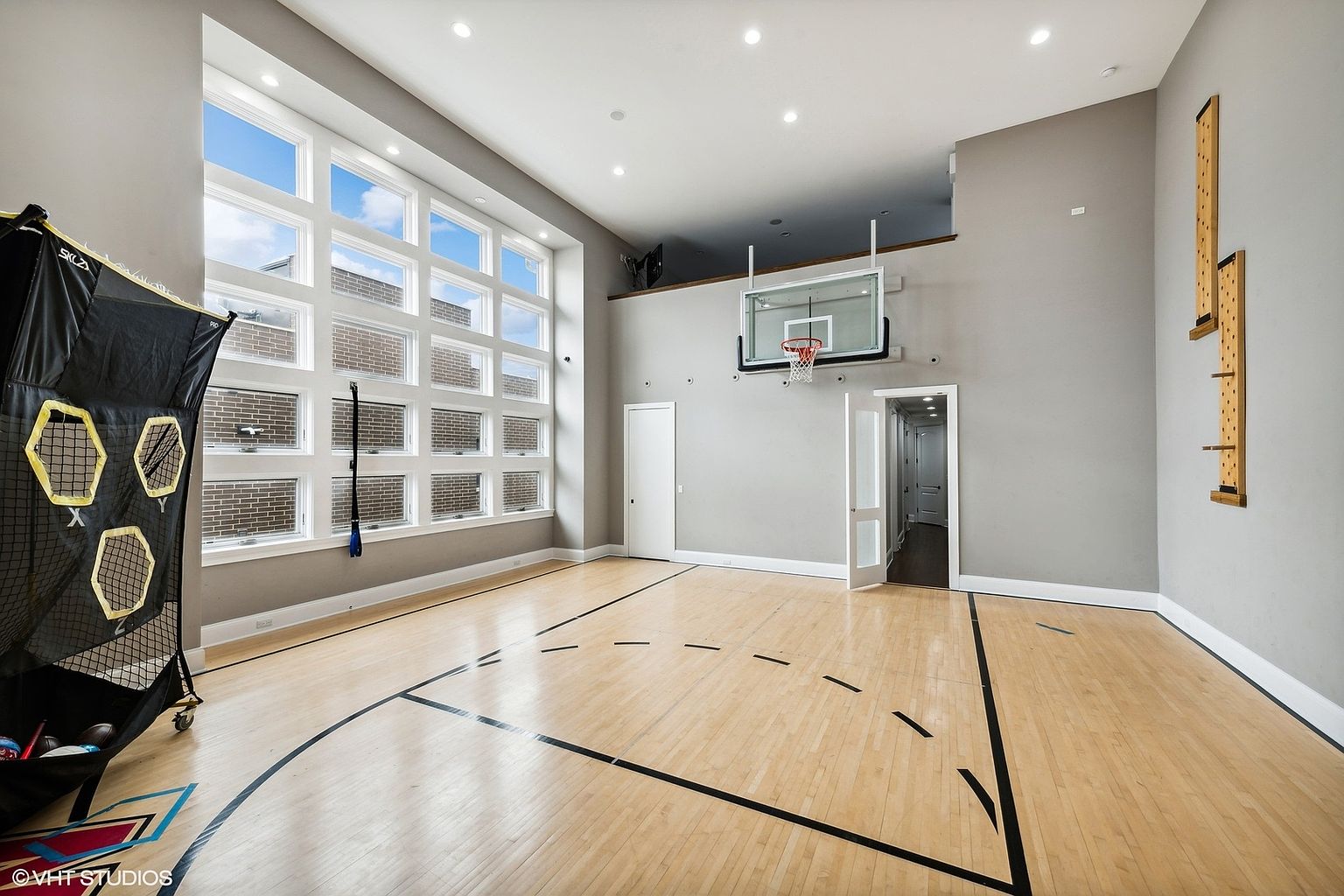 This is an interior shot of a home gym featuring a basketball court. The court has a light wood floor with black lines marking the boundaries. A basketball hoop is mounted on the wall, and a large window provides natural light. The walls are painted in a neutral gray tone, and there are recessed lights in the ceiling.