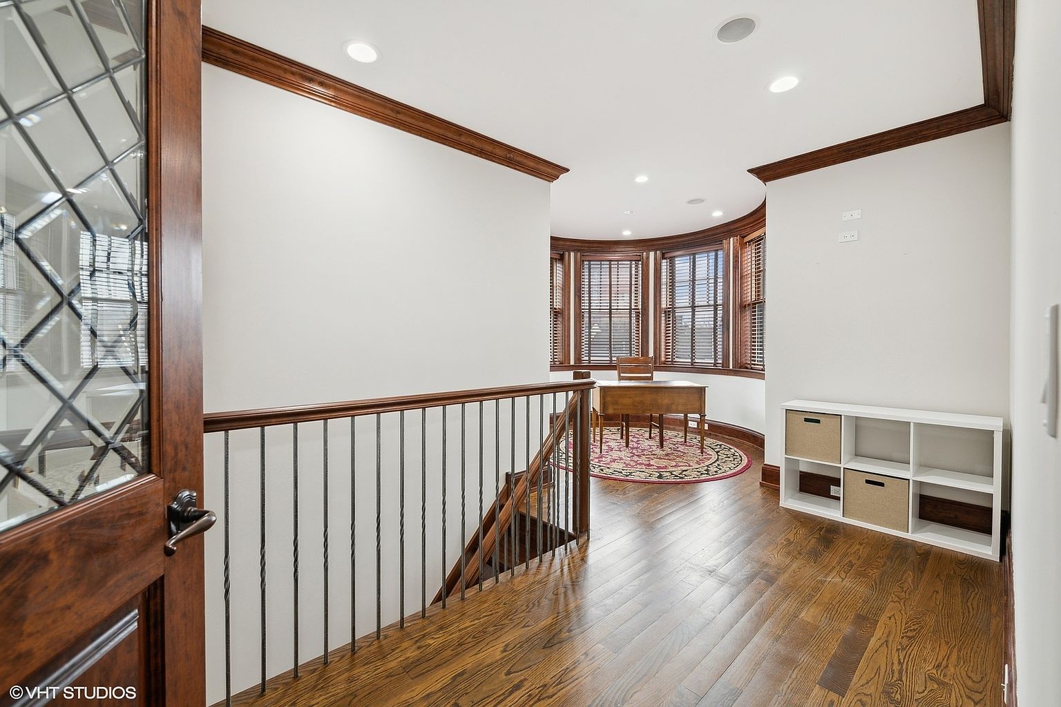 This is an interior shot of a hallway and stairs in a home. The hallway features hardwood floors, white walls with dark wood trim, and a staircase with wrought iron railings. A bay window area with a desk and chair is visible at the end of the hallway, and a white shelving unit is on the right. The perspective is from the top of the stairs looking into the hallway.