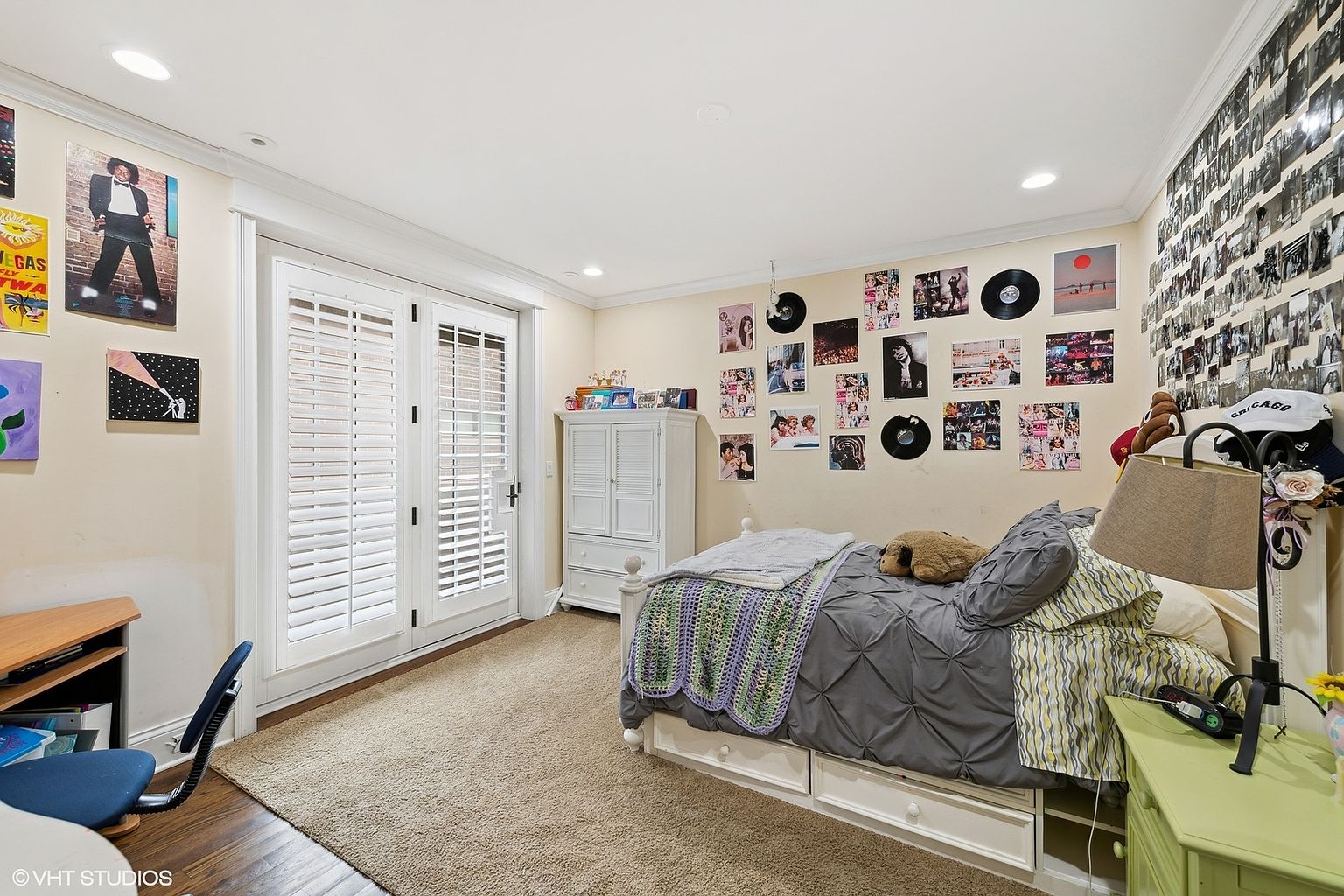 This is a cozy bedroom featuring a bed with a gray comforter and a patterned throw blanket. The walls are adorned with posters and records, creating a personalized and youthful atmosphere. A white dresser and a desk are also visible, providing storage and workspace.