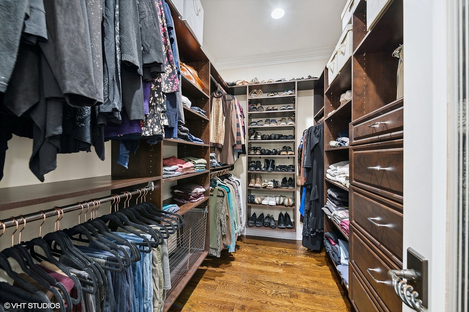 This is a well-organized walk-in closet featuring dark wood shelving and drawers. Clothes are neatly hung and folded, with a dedicated shoe rack in the center. The hardwood flooring adds a touch of elegance to the space, creating a luxurious and functional storage area.