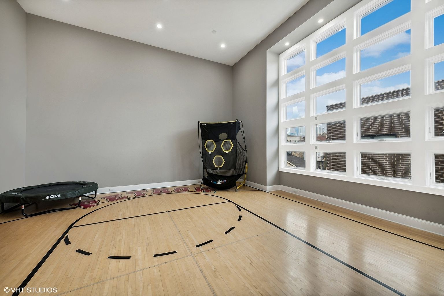 This is an interior shot of a home gym featuring a basketball court layout on a wooden floor. A large window provides natural light, and the room includes a trampoline and a basketball net. The walls are painted in a neutral gray tone, creating a clean and functional space.