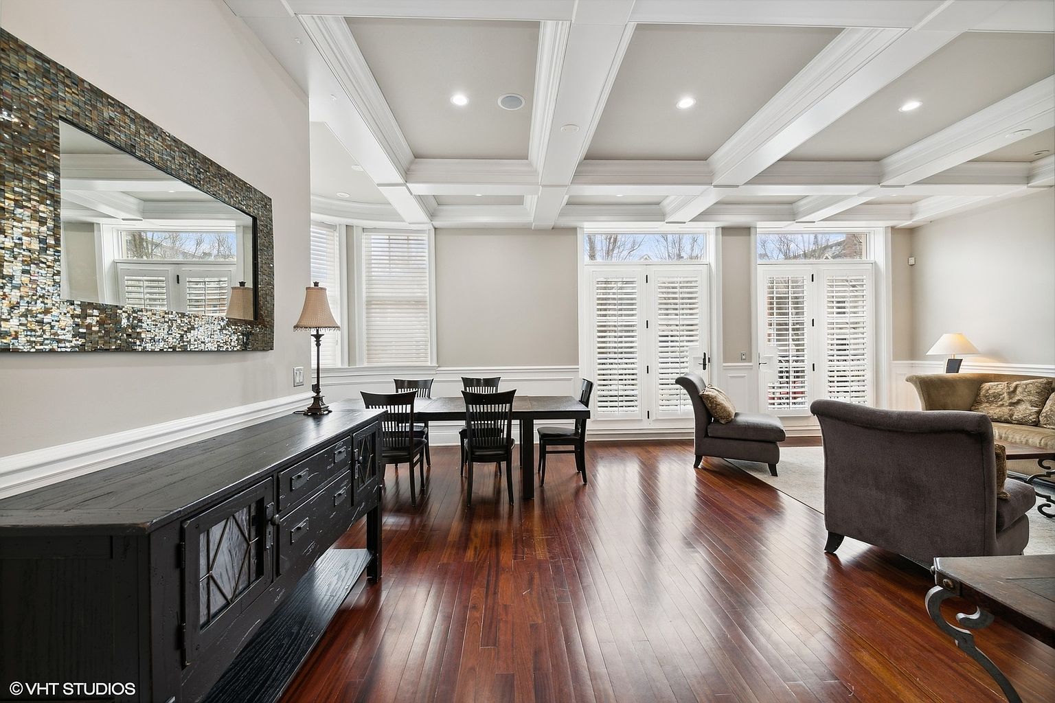 This is an interior shot of a dining room featuring dark hardwood floors and a coffered ceiling. A dark wood dining table with black chairs sits in the center of the room, complemented by a dark wood sideboard with a decorative mirror above it. Natural light streams in through the windows, enhancing the room's spacious feel.