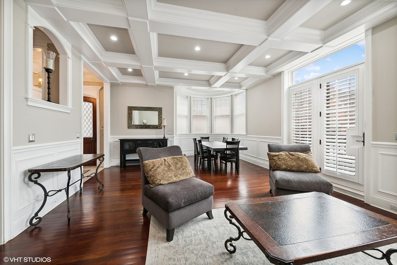 This is an interior shot of a living room featuring hardwood floors, coffered ceilings, and wainscoting. The room is furnished with two gray armchairs, a dark wood coffee table, and a console table. A dining area with a table and chairs is visible in the background, and natural light streams in through a large window with white shutters.