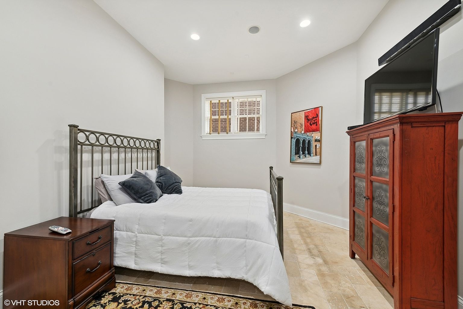 This is a cozy bedroom featuring a wrought iron bed frame with a white comforter and dark blue pillows. A dark wood nightstand sits beside the bed, and a matching cabinet with a TV on top is positioned against the opposite wall. The room is decorated with a framed art piece and a patterned rug, creating a warm and inviting atmosphere.