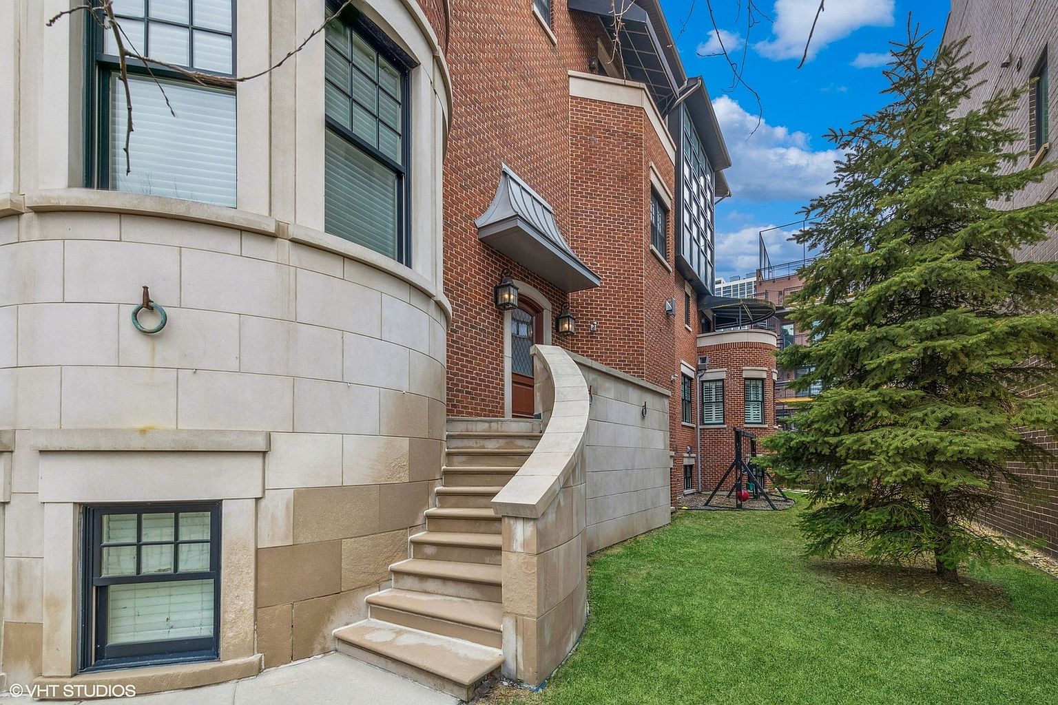 This image showcases the entryway of a brick and stone home. A curved stone staircase leads to a wooden front door with a decorative awning. The surrounding landscaping includes a well-maintained lawn and a mature evergreen tree, adding to the property's curb appeal.