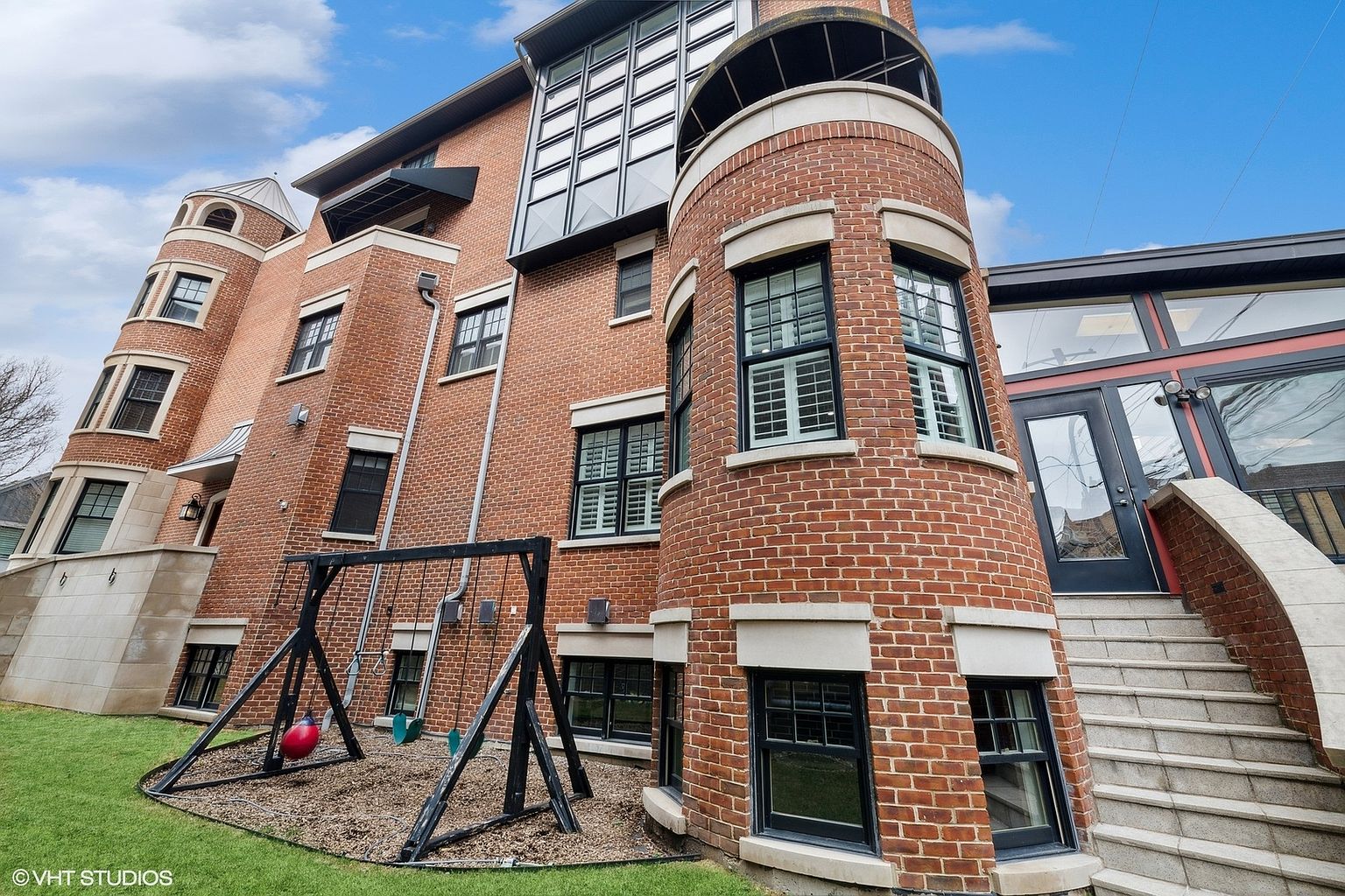 This is a side view of a unique brick building featuring a turret-like structure and modern architectural elements. The exterior includes a swing set in the yard, adding a playful touch to the property. The combination of traditional brickwork and contemporary design creates an interesting and appealing aesthetic.