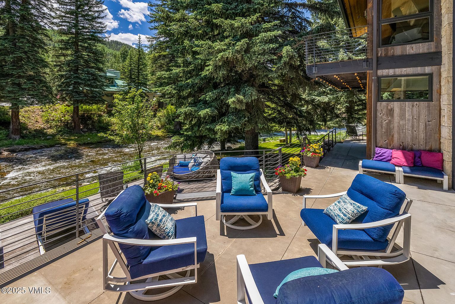This spacious outdoor deck features a comfortable seating area with blue cushioned chairs and a matching ottoman, perfect for relaxing while overlooking a scenic river. The deck is framed by a metal railing and surrounded by lush pine trees, creating a serene and private atmosphere. A secondary seating area with vibrant pink and purple pillows is visible along the side of the home, which is finished with natural wood siding.