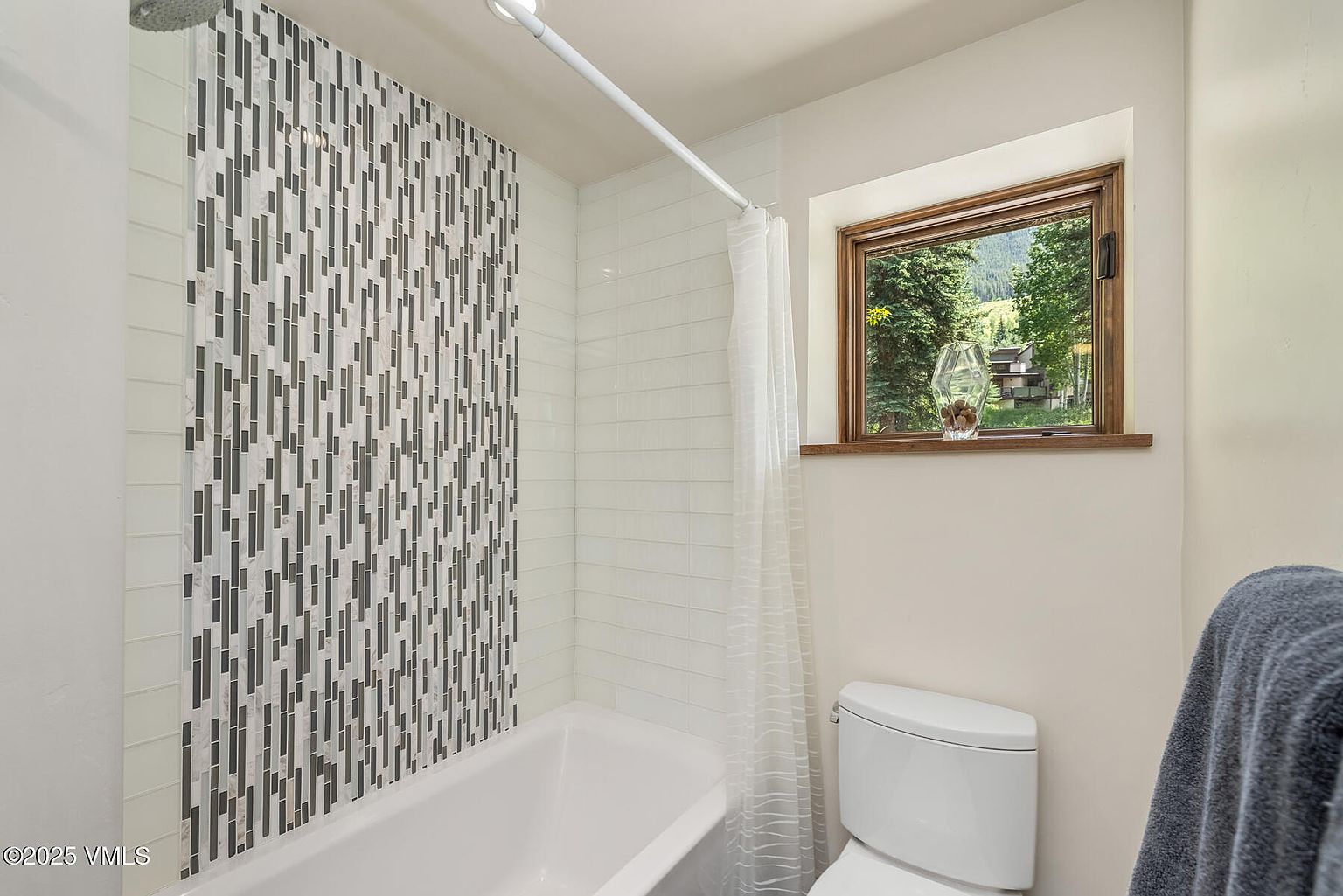 This bathroom features a shower-tub combination with a striking vertical accent wall of patterned, multi-toned tiles flanked by clean white subway tiles. A white toilet sits adjacent to the tub, and a window with a wood frame provides a view of the surrounding trees and landscape. The space is bright and functional, with a simple shower curtain rod and a decorative glass vase on the window sill.