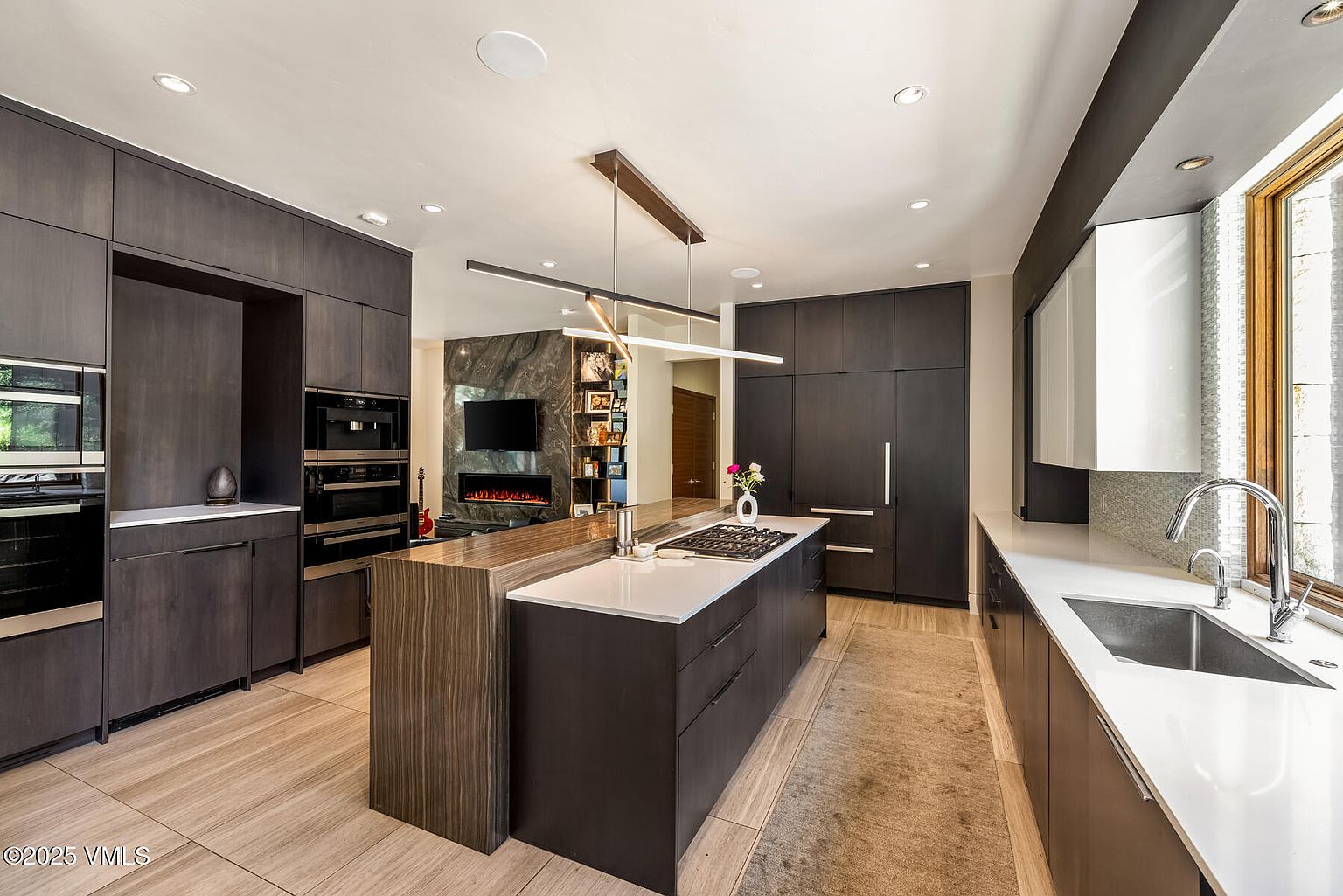 This sleek, contemporary kitchen features dark cabinetry paired with a large central island topped with a light-colored stone surface and an integrated gas cooktop. A linear pendant light fixture hangs above the island, while the adjacent wall showcases a tall, dark stone-clad chimney breast surrounding a fireplace and television. The space is finished with light-toned wood flooring and a long, minimalist sink area, creating a sophisticated and cohesive open-concept design.