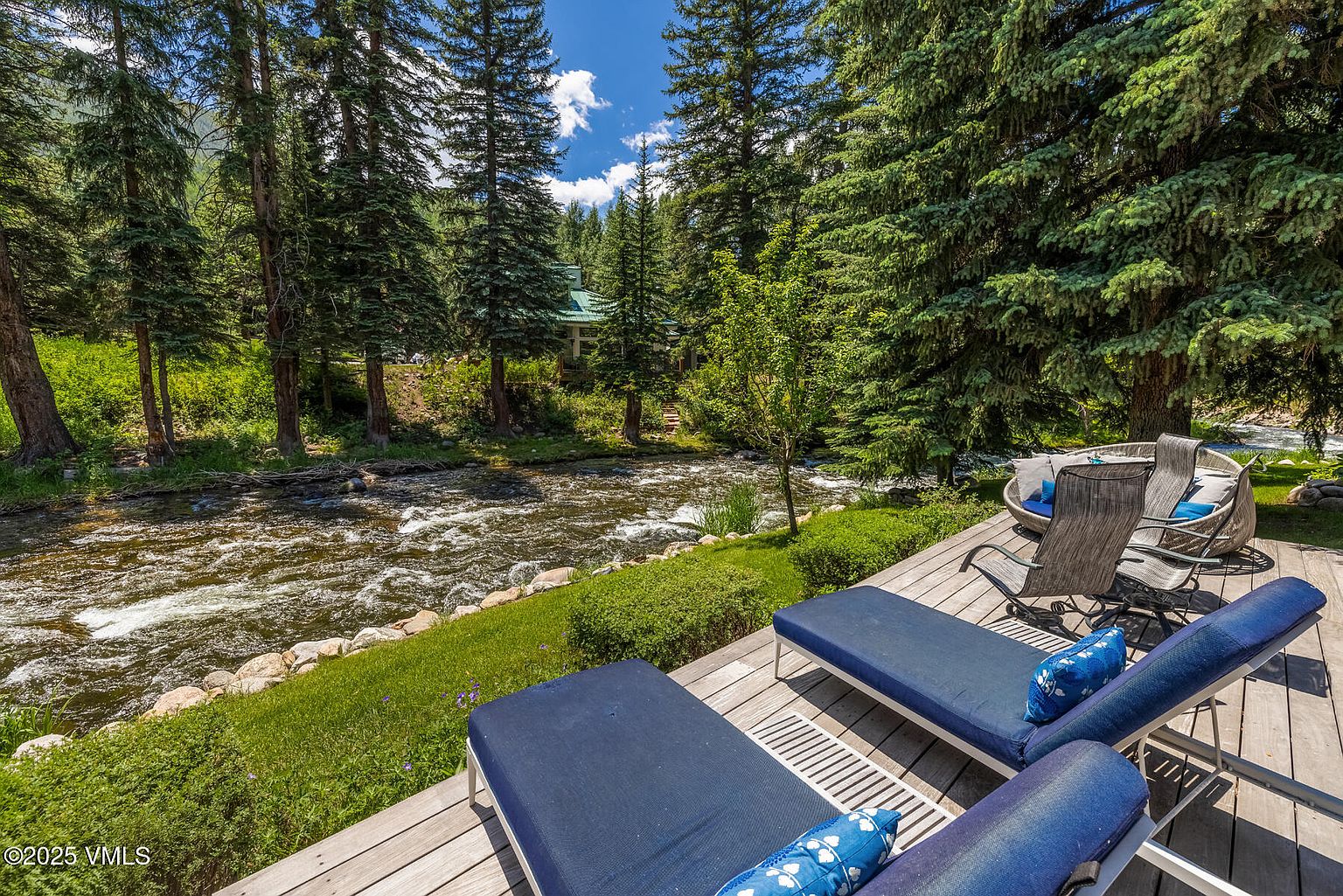 This serene outdoor deck offers a tranquil retreat overlooking a rushing mountain stream. The space is furnished with comfortable lounge chairs and a circular seating area, perfect for enjoying the natural surroundings and lush forest backdrop. The perspective captures the peaceful integration of the wooden deck with the vibrant green landscape and the flowing water.