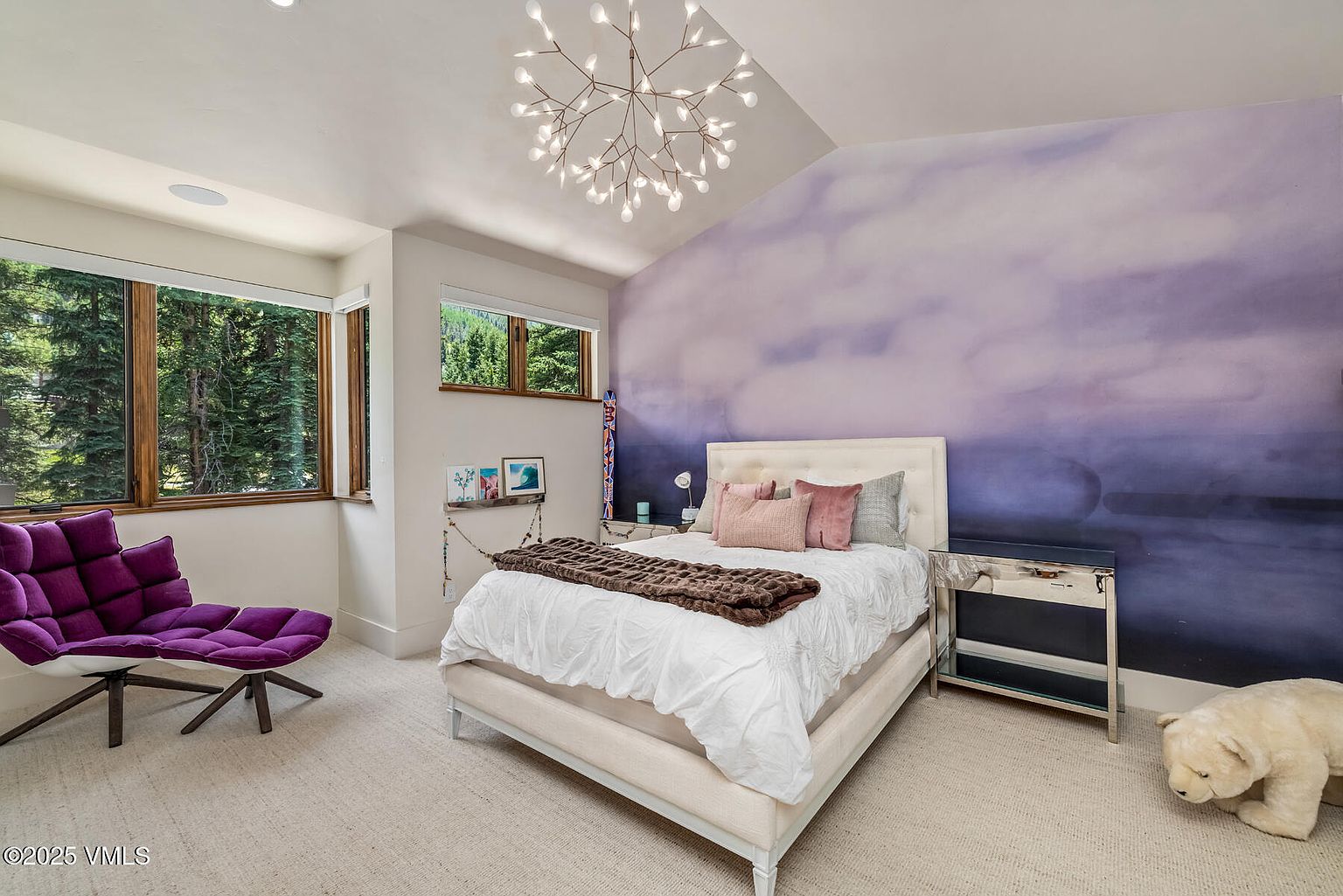 This bedroom features a striking purple ombre accent wall that serves as a focal point behind a light-colored upholstered bed. The room is illuminated by a modern, branching chandelier and includes a plush purple lounge chair, a mirrored nightstand, and a whimsical polar bear statue. Large windows provide a view of the surrounding forest, creating a serene and playful atmosphere.