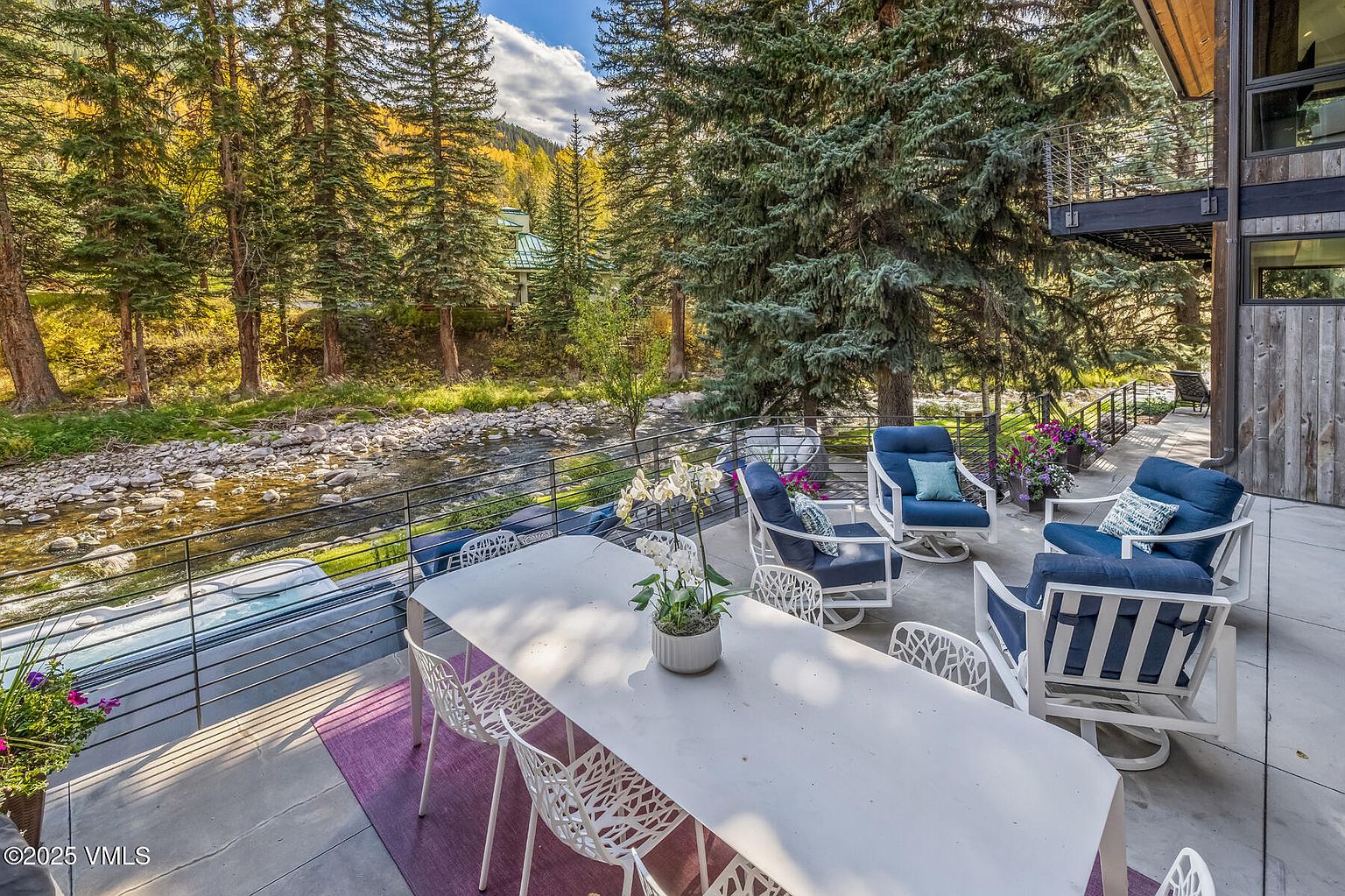 This spacious outdoor deck offers a serene retreat overlooking a rushing mountain stream and a lush, wooded landscape. The area is furnished with a modern dining set and comfortable lounge chairs, all set upon a clean concrete surface with a purple area rug. The scene captures a peaceful, high-end mountain lifestyle, framed by tall evergreen trees and a contemporary home exterior.