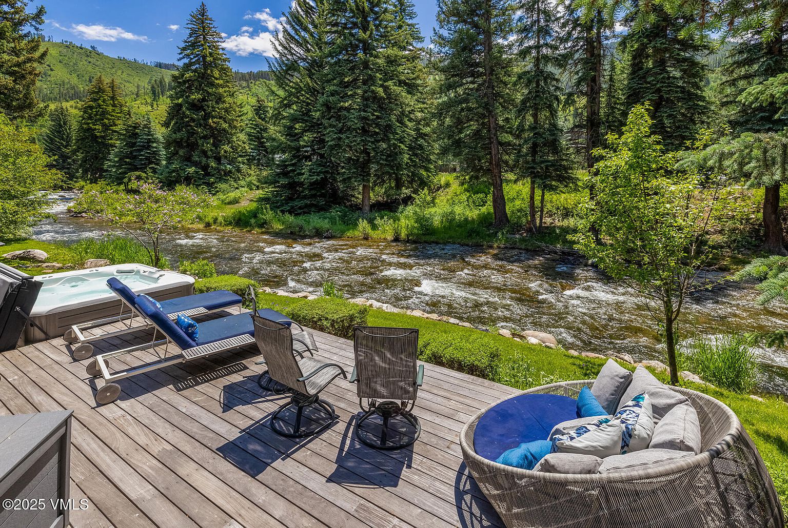 This expansive wooden deck offers a serene outdoor living space overlooking a rushing mountain river. The area is furnished with two blue lounge chairs, two swivel chairs, and a large, circular wicker daybed with plush cushions, all positioned to enjoy the scenic waterfront view. A hot tub is tucked into the corner of the deck, completing this tranquil retreat surrounded by lush pine trees and natural landscape.