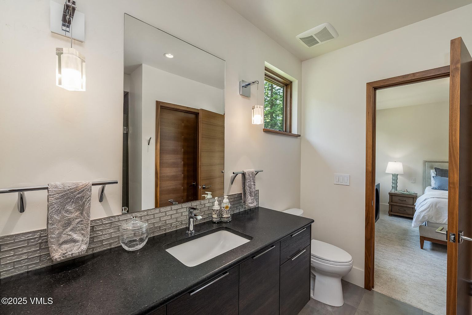 This modern bathroom features a dark vanity with a stone countertop and a rectangular undermount sink, complemented by a textured tile backsplash. A large mirror spans the wall above the vanity, flanked by contemporary pendant lights, while a doorway provides a view into an adjacent bedroom. The space is finished with neutral walls and clean, minimalist fixtures, creating a sleek and functional aesthetic.