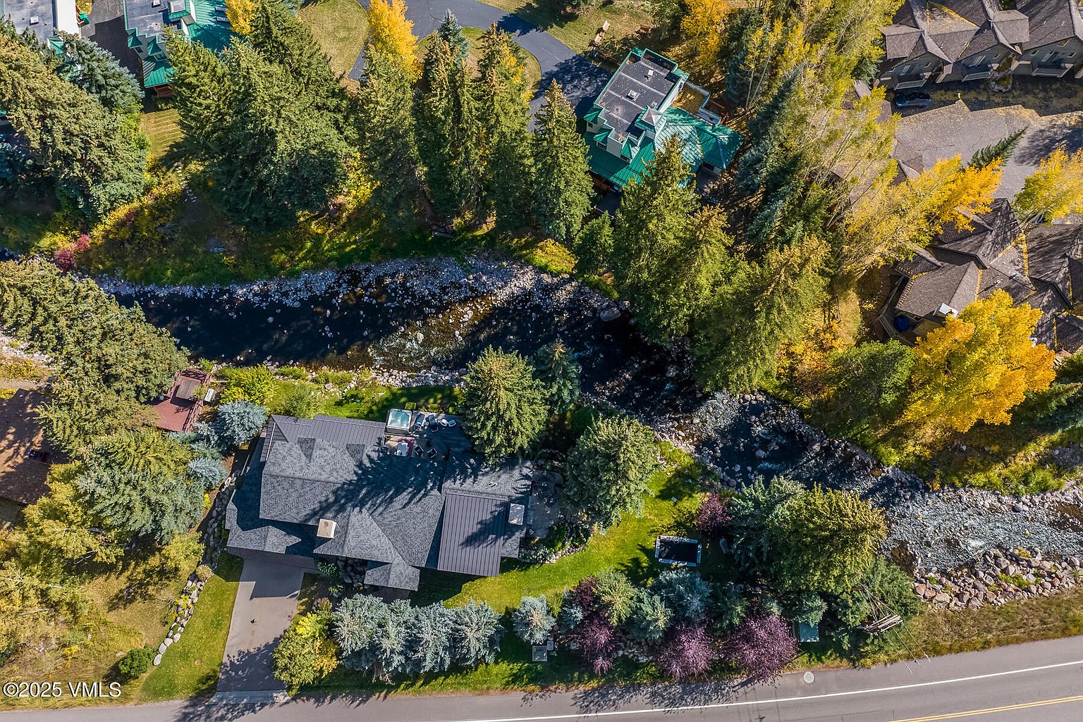 This high-angle aerial perspective captures a secluded residential property nestled in a lush, wooded landscape alongside a winding river. The home features a dark-toned roof and a multi-level deck area, surrounded by mature evergreen and deciduous trees that display vibrant autumn colors. A paved driveway leads to the residence, which is situated in a serene, natural setting with a small stream running through the grounds.