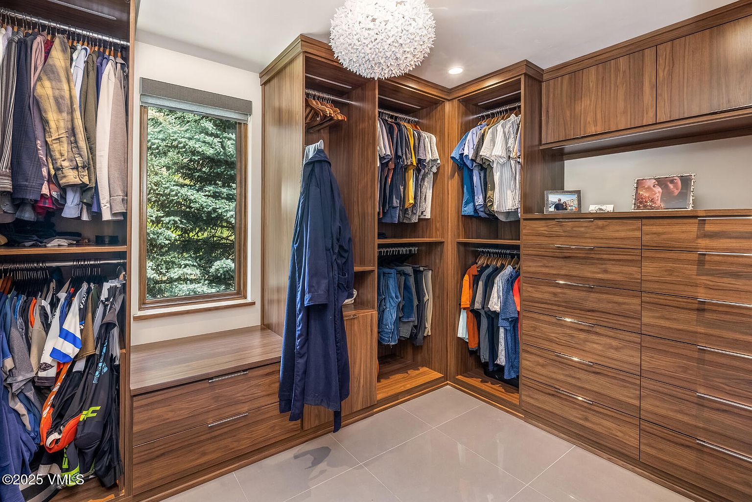 This spacious walk-in closet features custom dark wood cabinetry with extensive hanging space, open shelving, and a large bank of drawers. A window with a view of trees provides natural light, while a unique textured pendant light fixture hangs from the center of the room. The space is finished with neutral tile flooring and offers a clean, organized aesthetic suitable for a primary suite.
