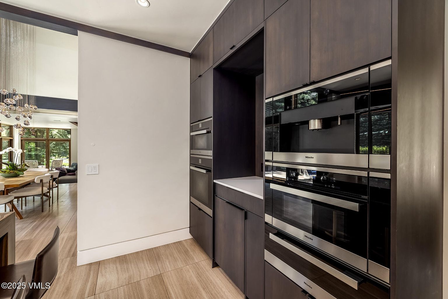 This modern kitchen area features a sleek, dark-toned cabinetry wall integrated with a suite of high-end Miele stainless steel appliances, including a built-in coffee system and ovens. A clean, light-toned countertop provides a workspace, while the adjacent open-plan dining area is visible through a wide doorway. The space is characterized by a sophisticated, minimalist aesthetic with warm wood-look flooring and a bright, airy transition to the living space.