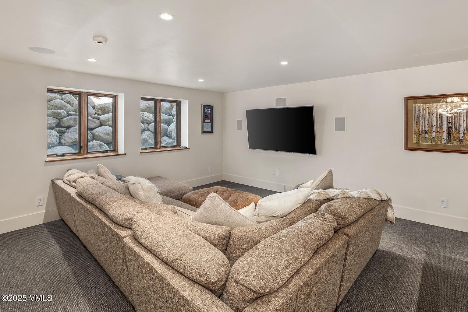 This cozy basement living room features a large, plush sectional sofa arranged around a central leather ottoman, creating an inviting space for relaxation. The room is equipped with a wall-mounted television and integrated speakers, while two windows look out onto a natural stone retaining wall. The neutral color palette and soft carpeting contribute to a comfortable, relaxed atmosphere.
