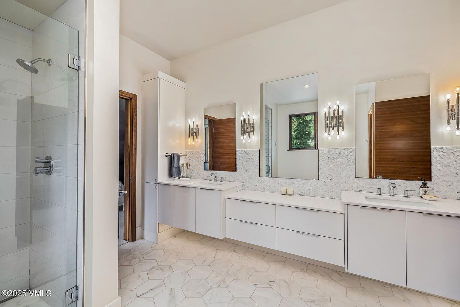 This primary bathroom features a sleek, modern design with a double vanity, white cabinetry, and a light-toned stone countertop. The space is accented by a hexagonal patterned tile floor and a white tiled shower enclosure with a glass door, while wall-mounted light fixtures flank the mirrors to create a bright, polished atmosphere. The perspective captures the full length of the vanity area, emphasizing the clean lines and contemporary aesthetic of the room.