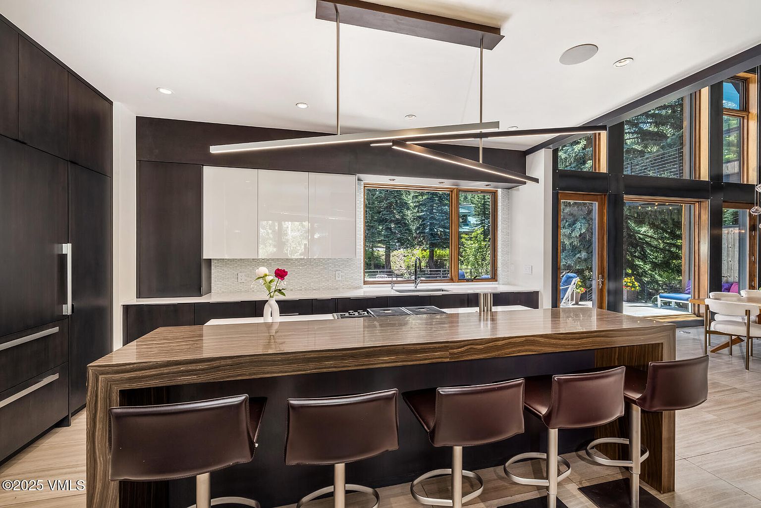 This modern kitchen features a large island with a prominent wood-grain countertop and integrated seating for five. The space is defined by a striking linear pendant light fixture, dark cabinetry, and a contrasting white upper cabinet section. Large windows provide a view of the surrounding trees, creating a bright and airy atmosphere that blends contemporary design with natural elements.