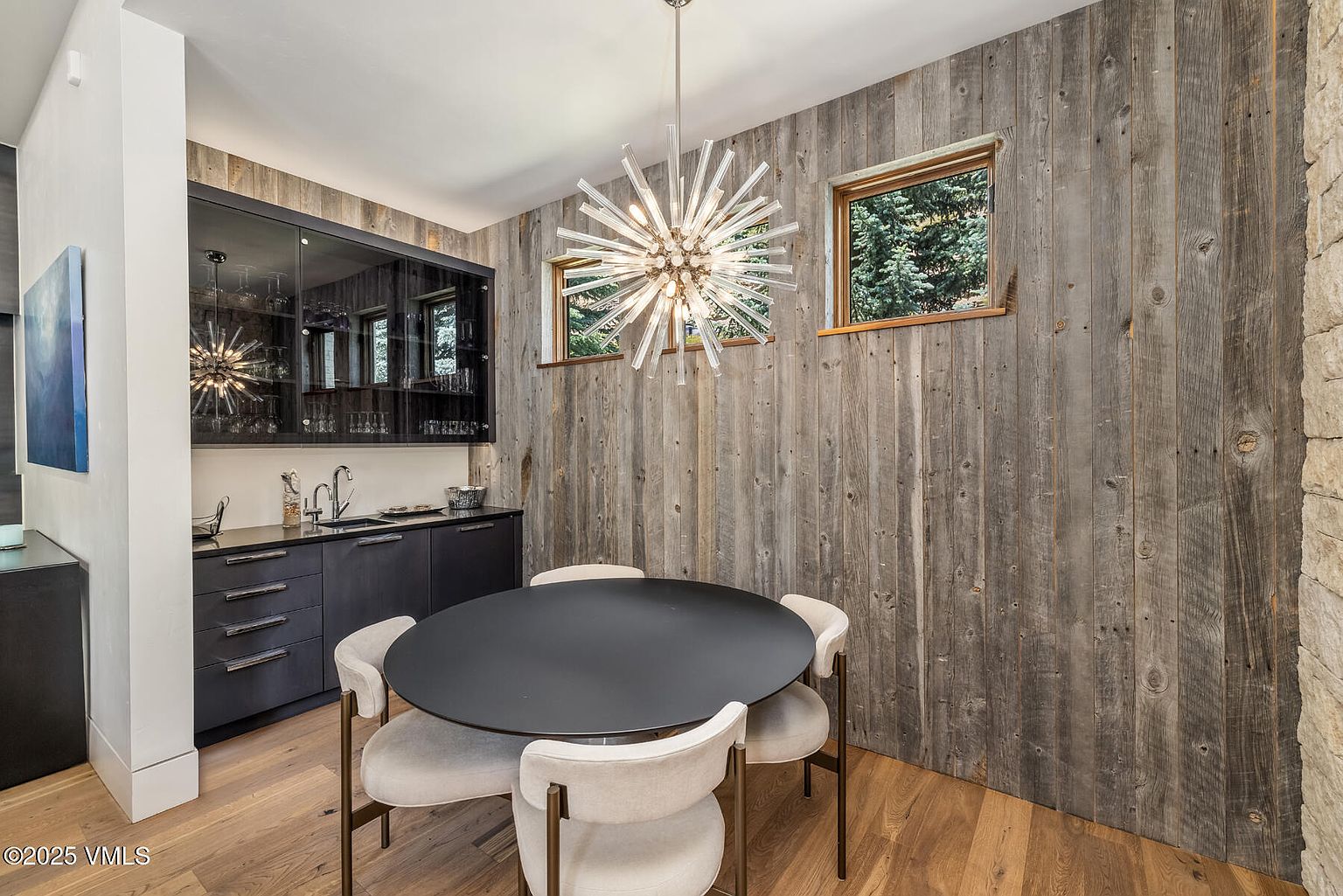 This dining area features a modern round table surrounded by four upholstered chairs, positioned beneath a striking starburst-style chandelier. The room is accented by a rustic wood-paneled wall with two windows and a built-in wet bar with dark cabinetry and glass-fronted upper cabinets. The space combines contemporary design elements with natural textures, creating a sophisticated and inviting atmosphere.