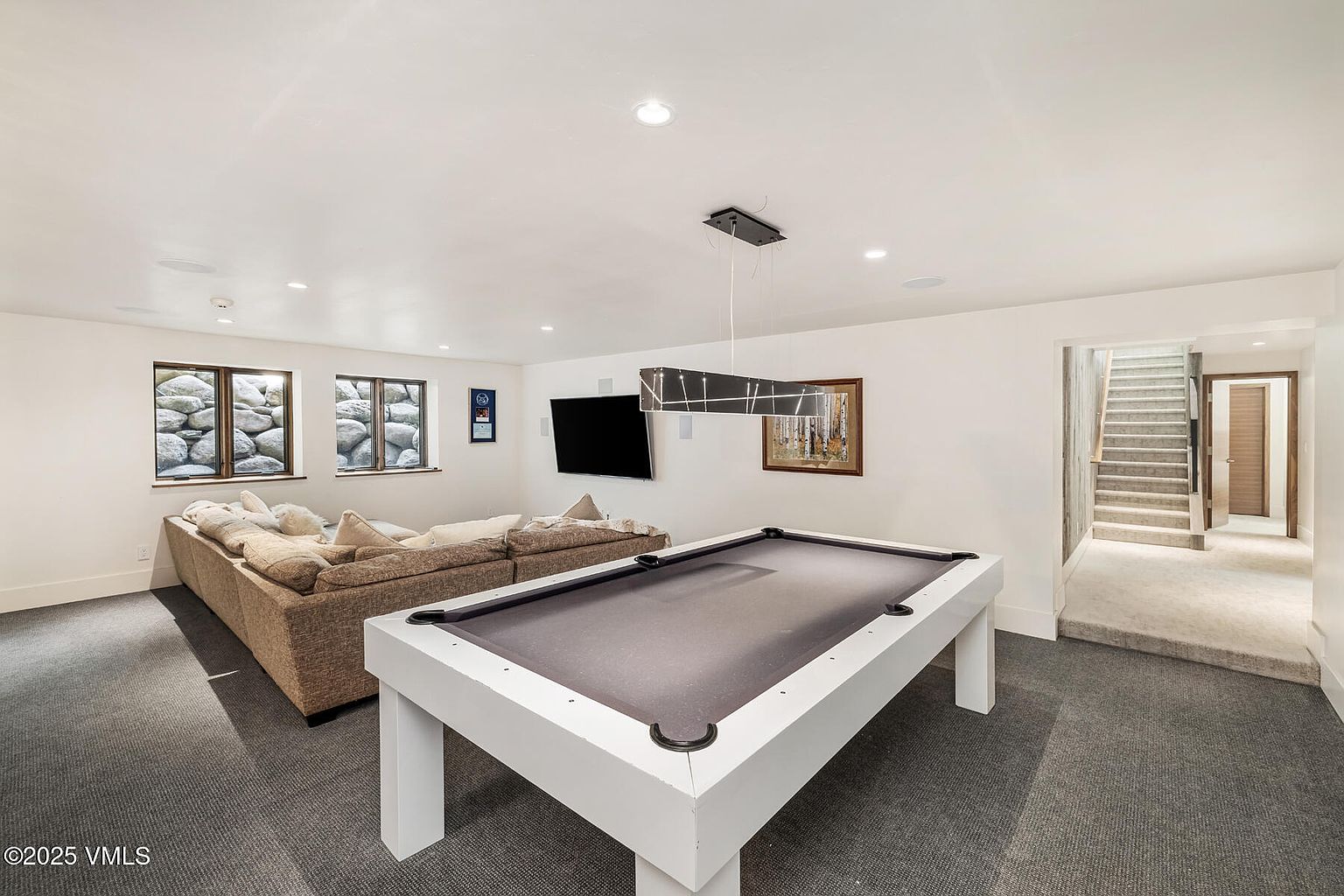 This spacious basement recreation room features a modern white pool table as the central focal point, positioned beneath a contemporary linear pendant light. To the left, a large, plush sectional sofa faces a wall-mounted television, creating a comfortable lounge area. The room is finished with neutral walls, recessed lighting, and carpeted flooring, with a staircase leading to an upper level visible on the right.