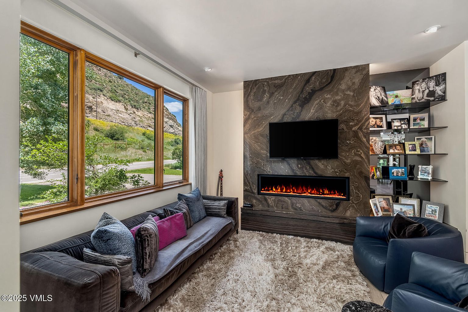 This contemporary living room features a striking, dark stone-clad chimney breast that houses a wide, linear electric fireplace and a wall-mounted television. A plush, textured area rug anchors the space, which is furnished with a long, dark sofa accented by vibrant purple and blue pillows, alongside a deep blue leather armchair. Large windows provide an expansive view of the natural landscape outside, filling the room with bright, natural light.