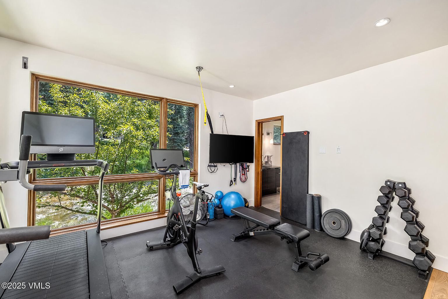This home gym features a dedicated workout space equipped with a treadmill, a stationary bike, a weight bench, and a rack of dumbbells. Large windows provide an expansive view of the lush greenery outside, filling the room with natural light. The space is finished with dark rubber flooring, creating a functional and professional atmosphere for exercise.