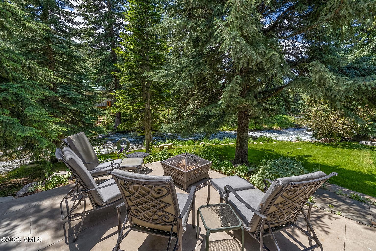 This serene outdoor patio space features a comfortable seating arrangement with four cushioned chairs and a central metal fire pit, perfect for relaxing by the water. The patio overlooks a lush, green lawn and a rushing river, framed by mature evergreen trees that provide natural shade and privacy. The perspective is a slightly elevated eye-level shot, capturing the tranquil connection between the private living space and the natural landscape.