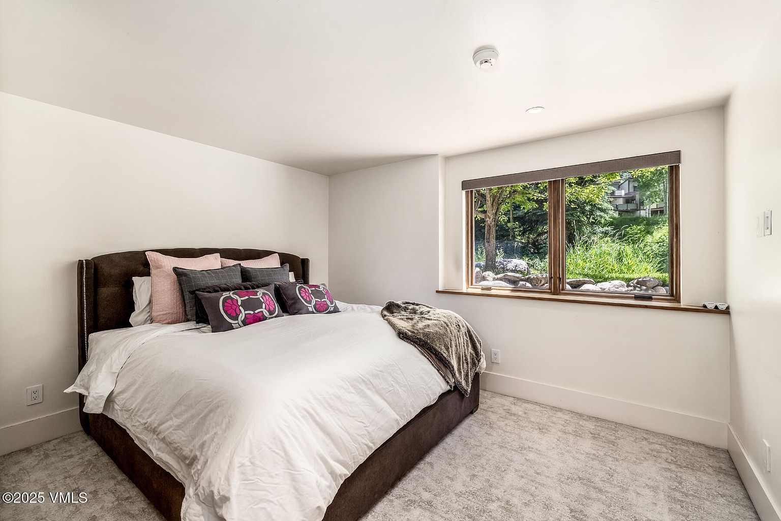 This cozy bedroom features a plush, dark-toned upholstered bed frame centered against a clean, white-walled backdrop. A large window with a wooden frame provides a view of the lush greenery outside, allowing natural light to brighten the space. The room is finished with neutral carpeting and a simple, modern aesthetic, creating a peaceful and inviting atmosphere.